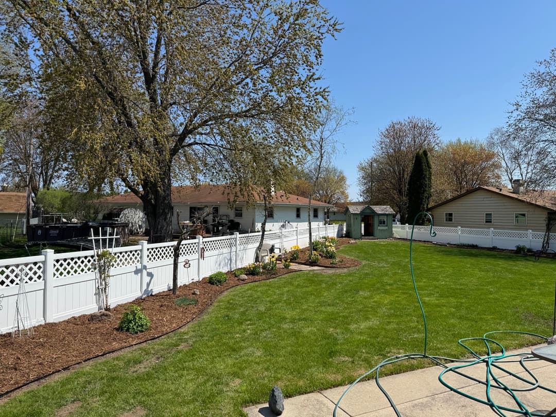 Lush backyard with green lawn, white fence, trees, and garden beds under clear blue sky.