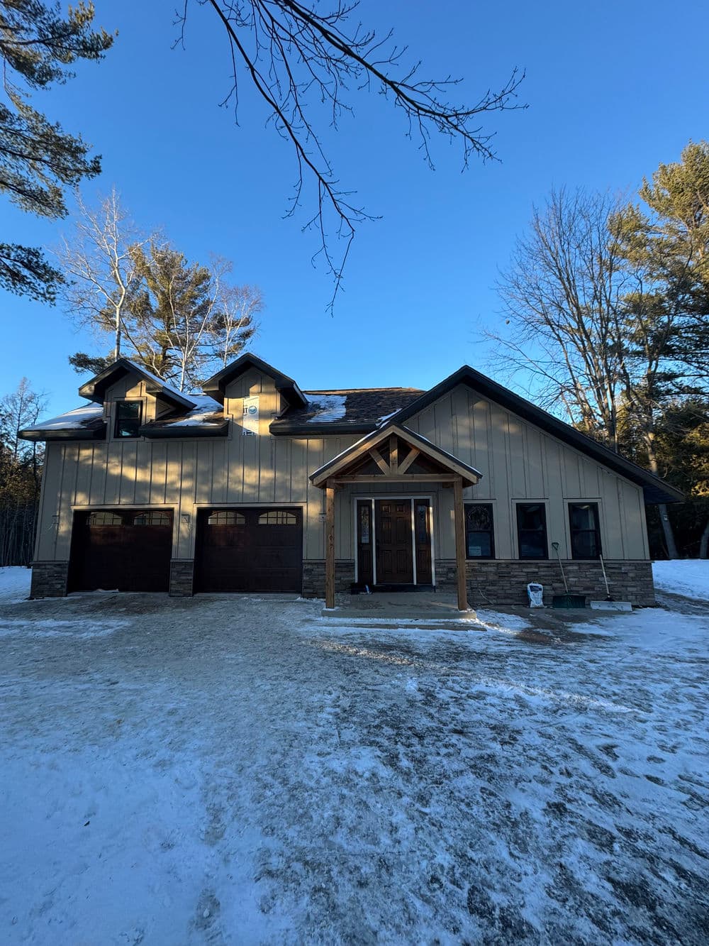 Modern house with a wooden porch, snow-covered yard, and two-car garage in winter setting.