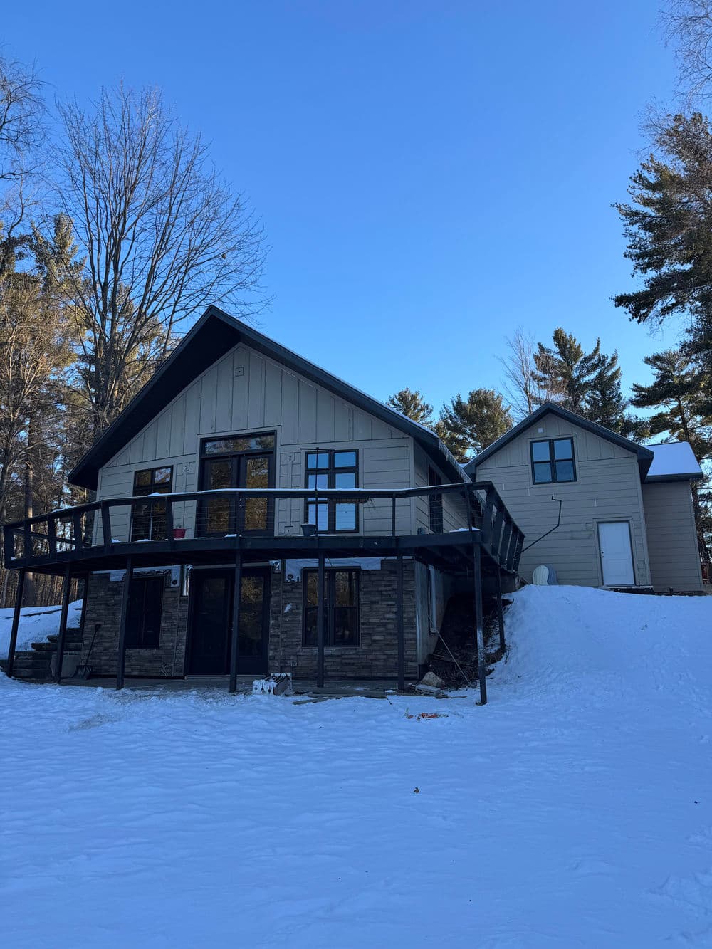 Modern two-story house with a deck, surrounded by snow and trees on a clear blue sky day.